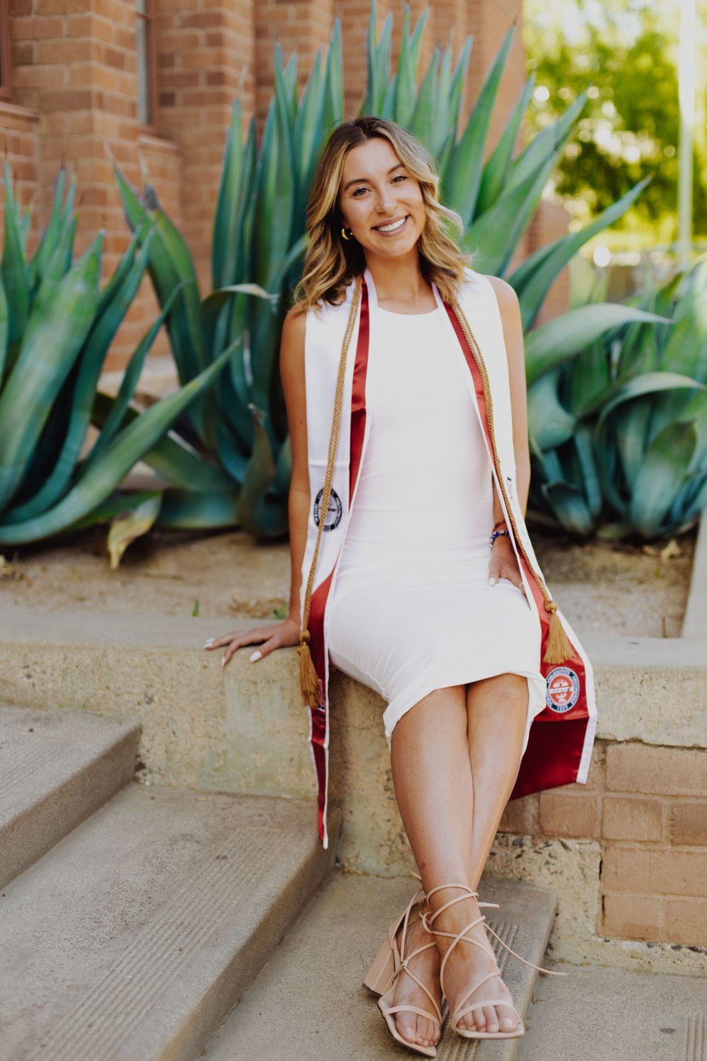 Graduate in white dress and red stole poses near desert plants on campus.