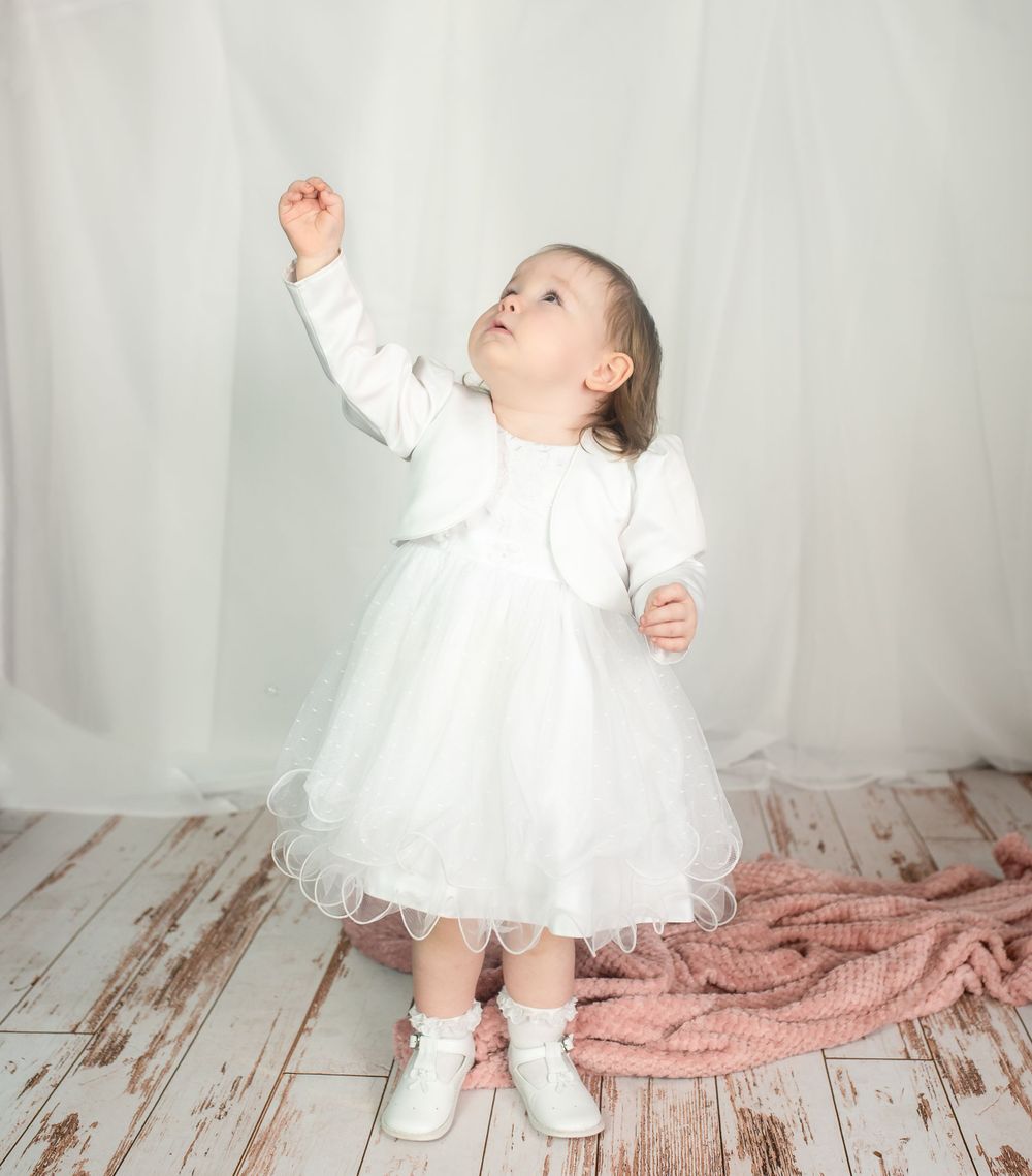 Child in white dress reaching upward while standing on wooden floor with pink blanket.