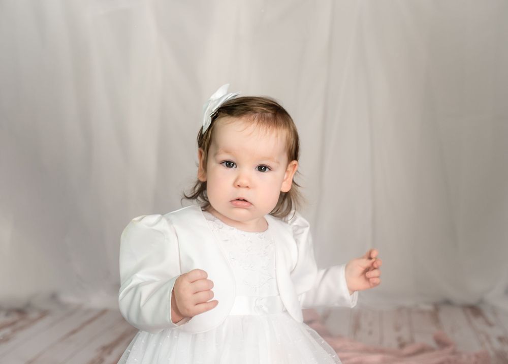 Small child in elegant white dress and bow against neutral background.