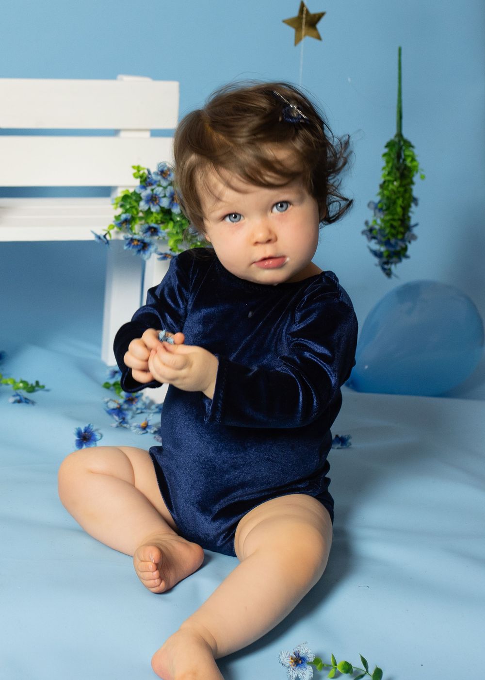 Baby in navy velvet outfit sitting on blue set with flowers and star decor.