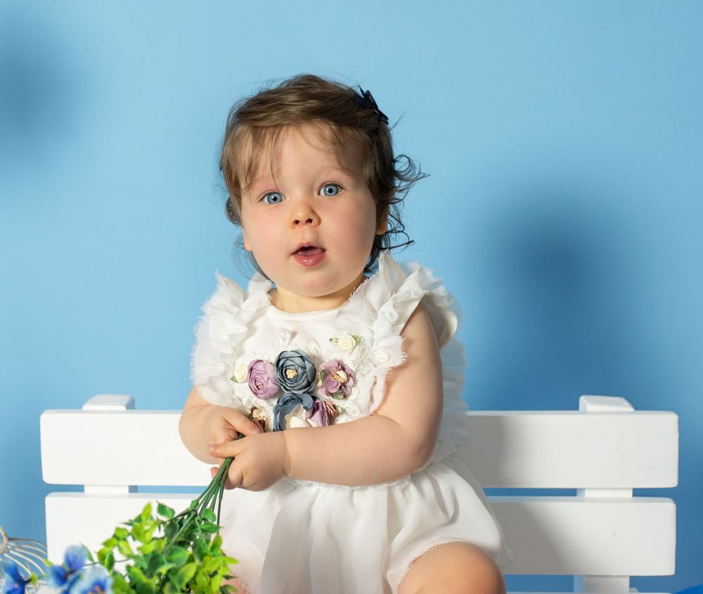 Child in white outfit sitting on white bench with greenery and blue background.