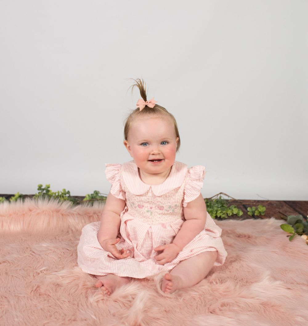 Baby wearing pink dress sitting on pink furry rug with greenery accents.