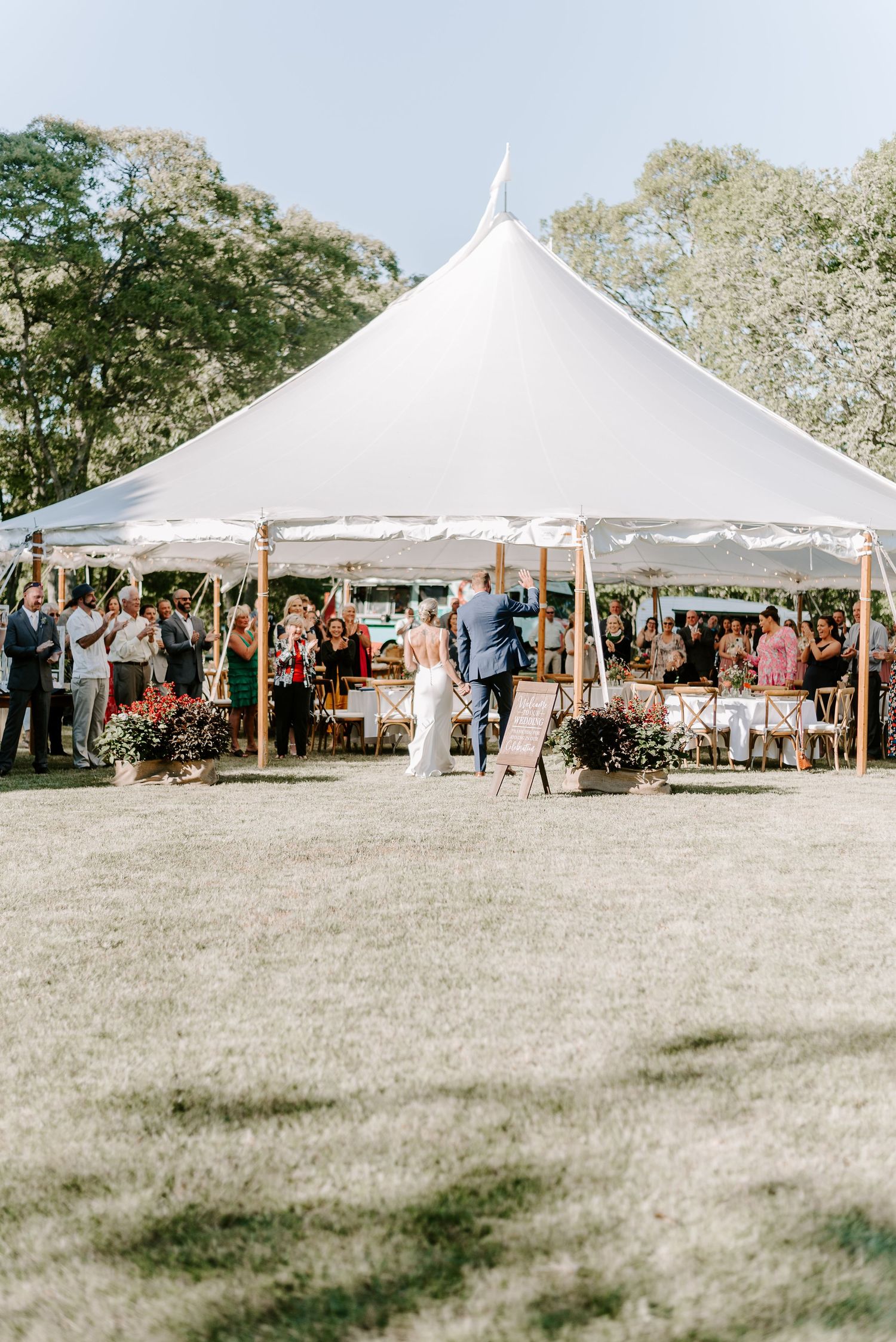 Large white tent wedding reception setup with guests gathered on a sunny outdoor lawn.