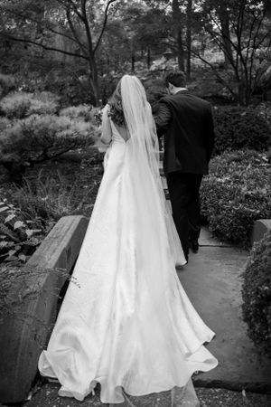 Black and white wedding photo showing bride's flowing train as couple walks garden path.