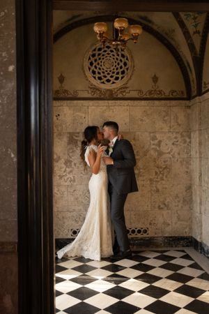 Romantic wedding portrait under ornate ceiling with checkerboard floor and vintage lighting.