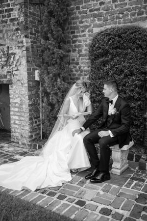 Black and white portrait of newlyweds seated by brick wall and manicured garden.