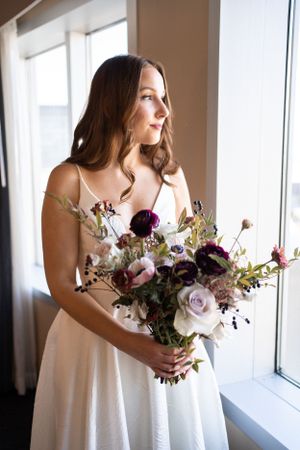 Bride holding romantic bouquet of purple and pink flowers by window in natural light.
