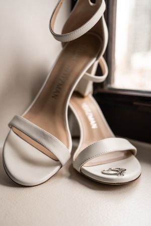 Elegant white bridal sandals with ankle strap displayed with wedding ring.