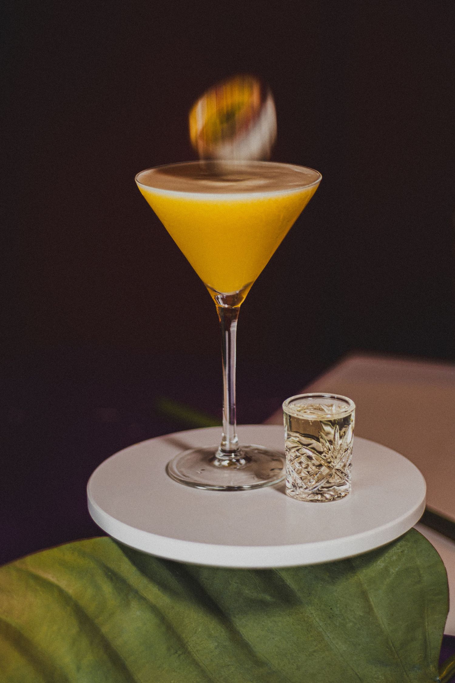 Passievrucht valt in een Pornstar Martini cocktail op een wit plankje, met een prosecco shot ernaast en een groen onderstuk.