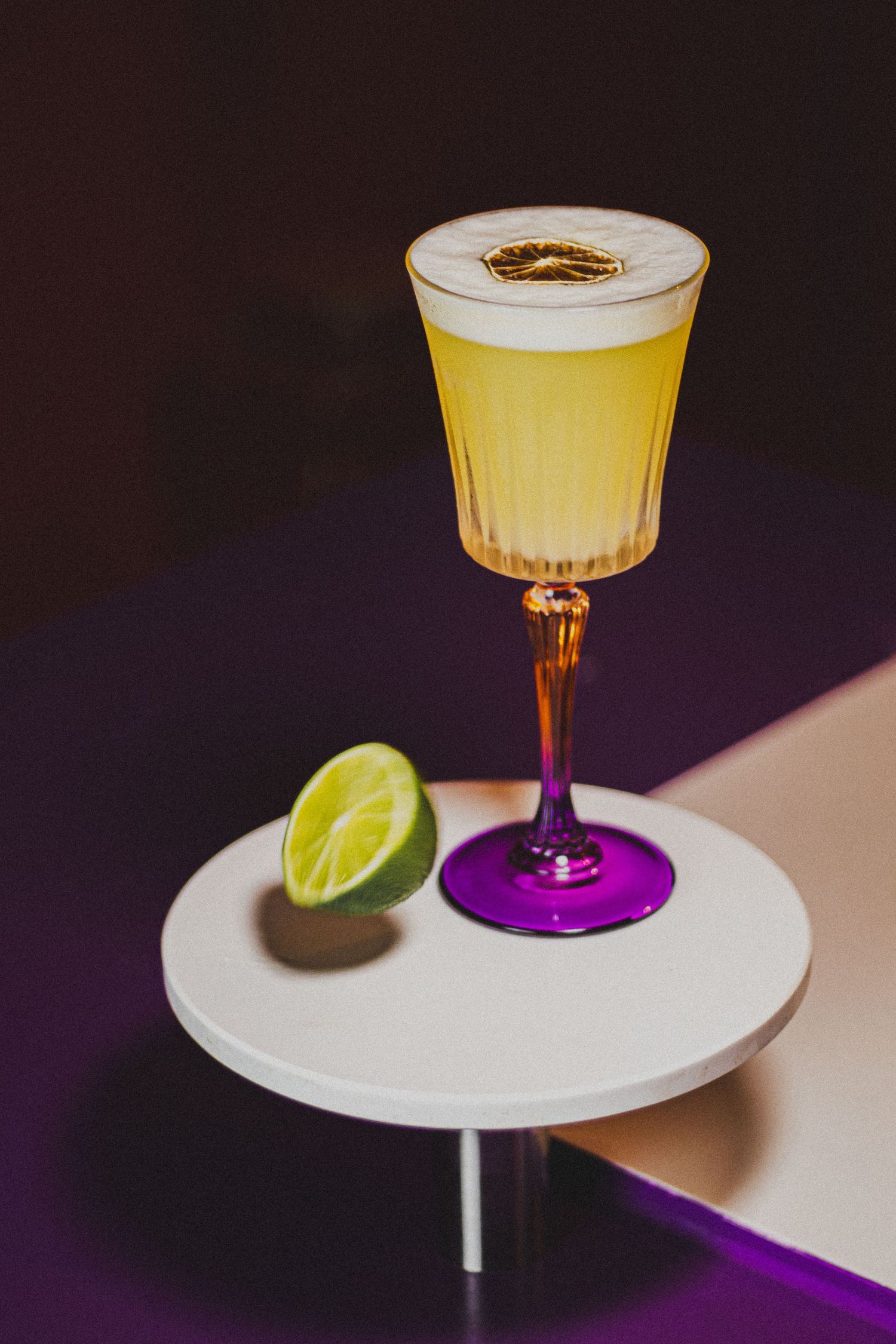 Stijlvolle gele cocktail op een paars voetglas met halve limoen op een modern wit tafeltje.