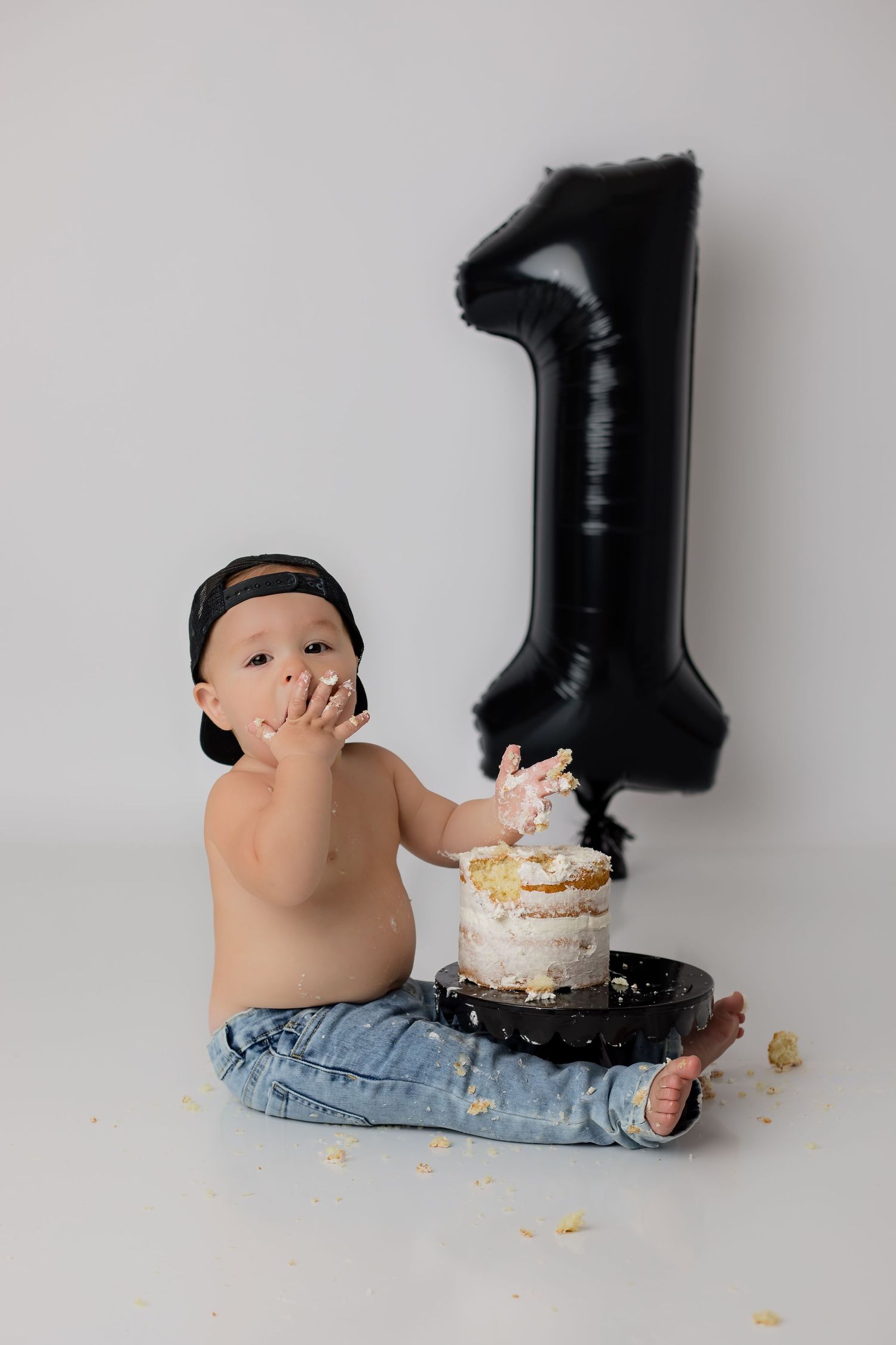 Simple white backdrop and a smiling baby boy eating cake in his jeans for his one year photoshoot.