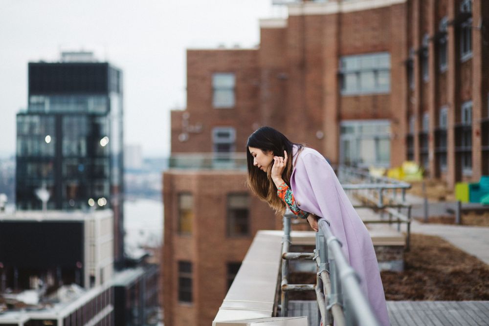 Person in lilac coat leaning over rooftop railing with cityscape view.
