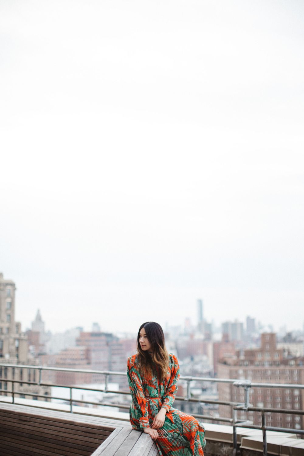 Person in floral dress sitting on wooden bench with Manhattan skyline in background.