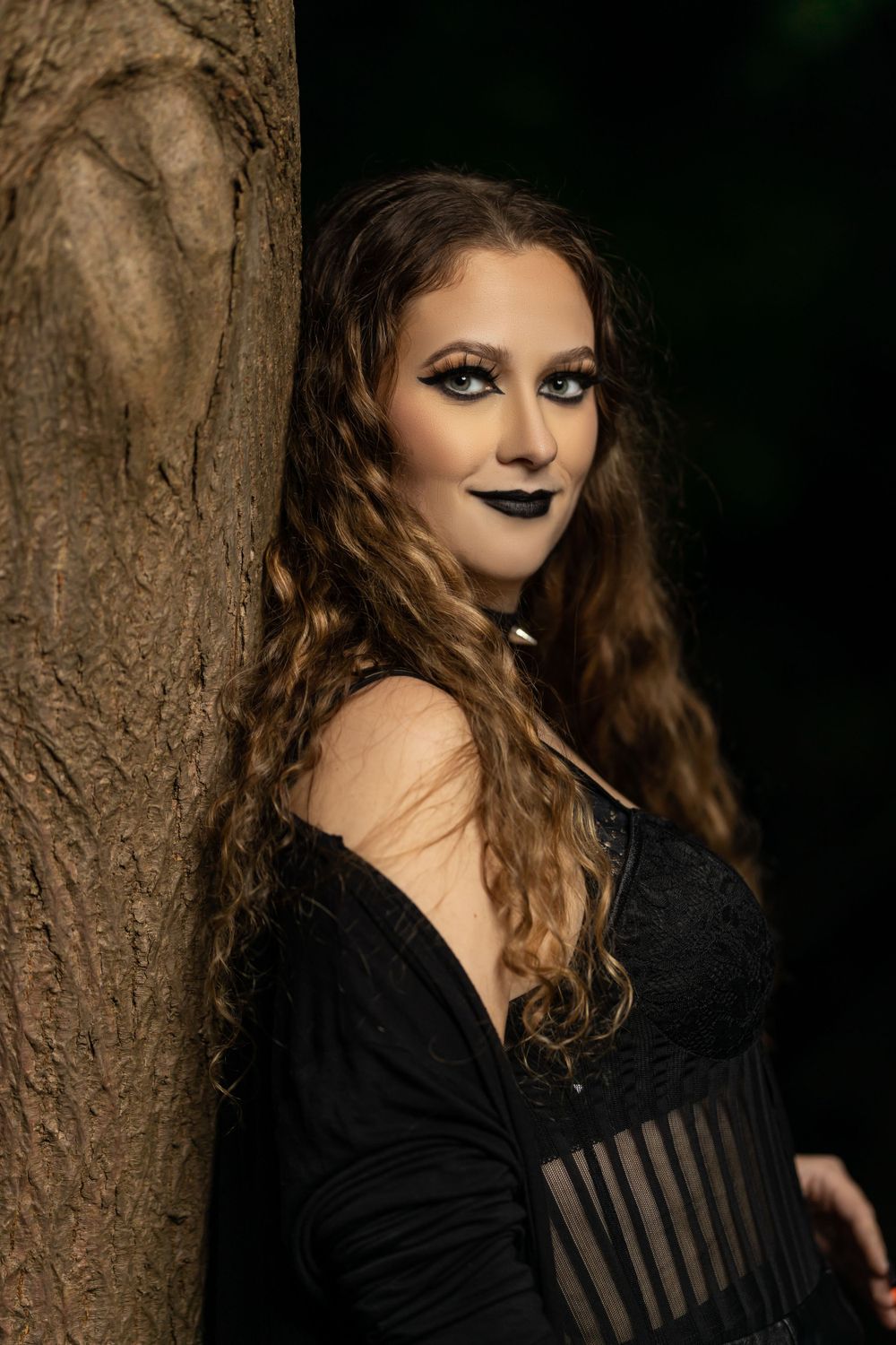 A dark gothic portrait showing someone in black attire leaning against a tree trunk.