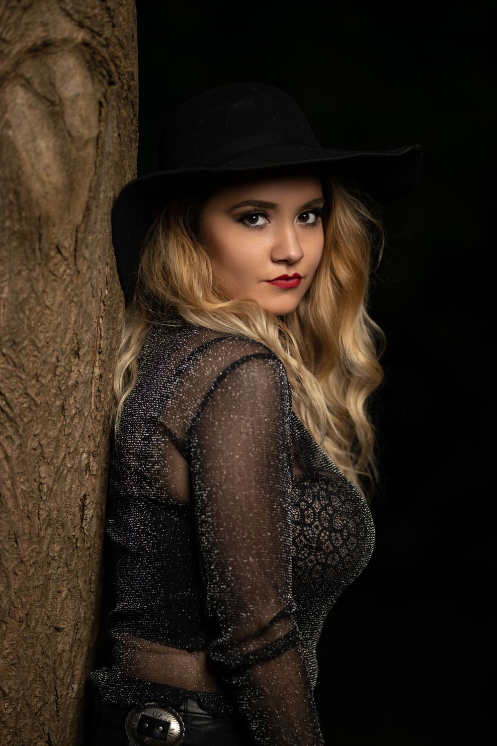 Dramatic portrait against tree trunk featuring dark hat and sheer lace top with moody lighting.