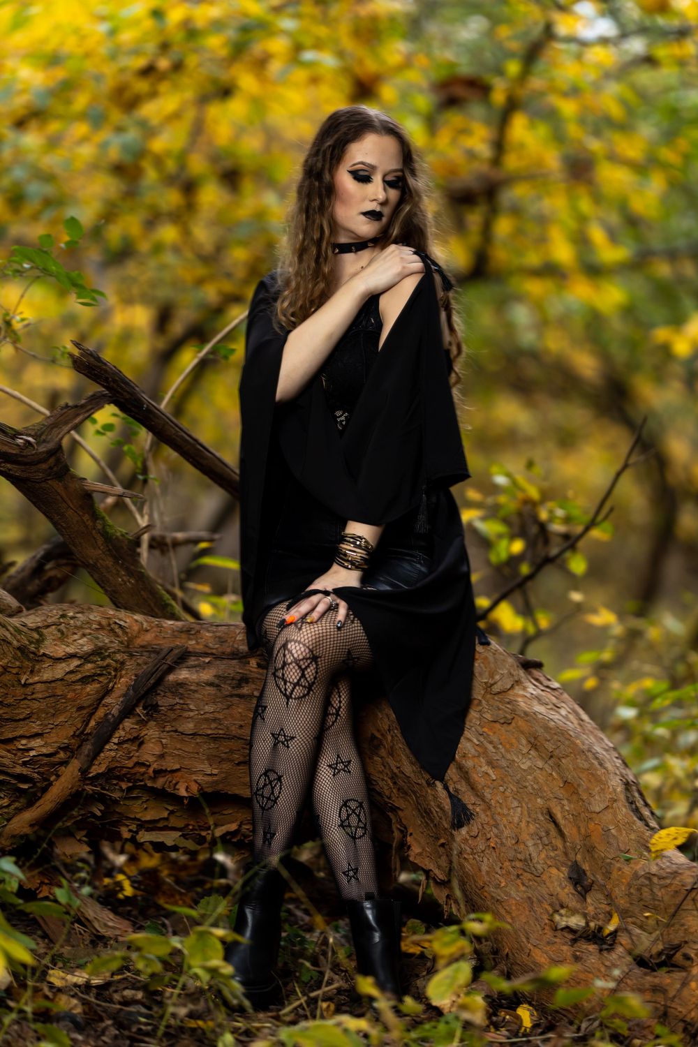 Gothic fashion portrait featuring black dress and patterned tights on rustic log setting.