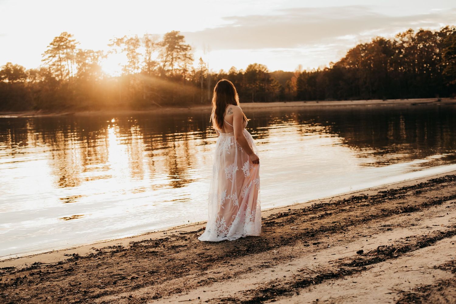A woman in a flowing floral dress stands by a serene lake at golden sunset surrounded by autumn trees.