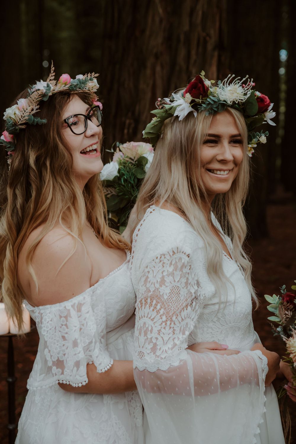 Lesbian brides in white lace dresses share a joyful moment in the woods.
