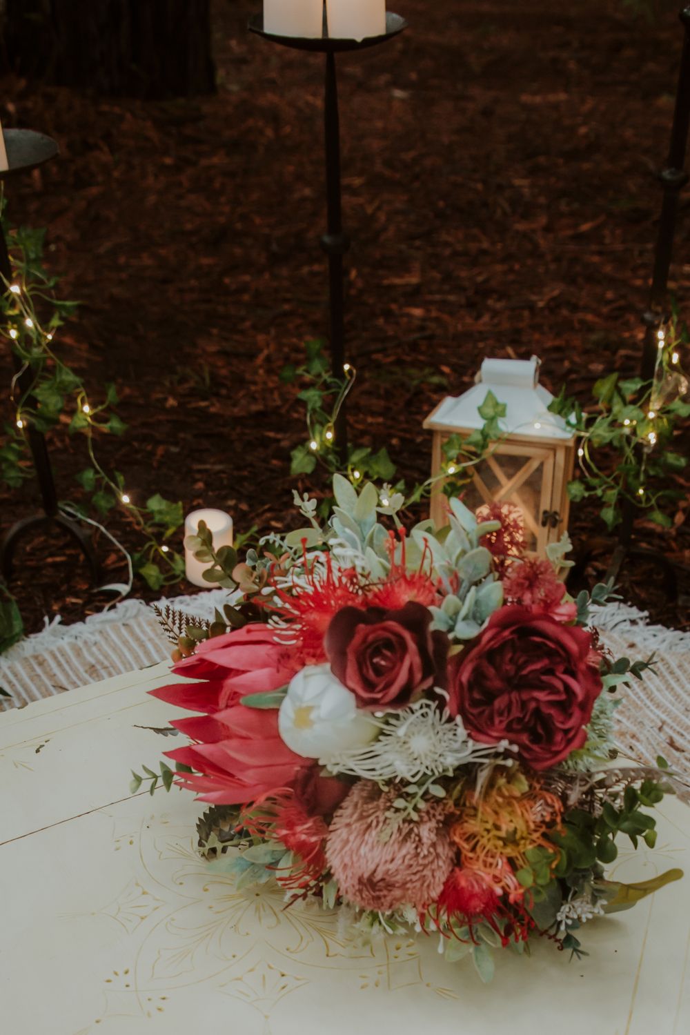 A vibrant bouquet with red roses, protea, and eucalyptus next to a white lantern on a table.