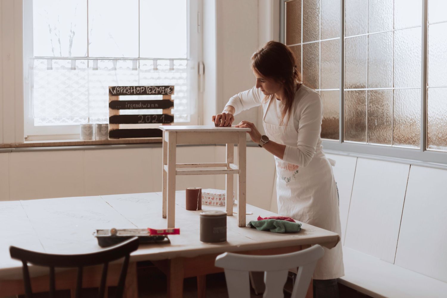 Jemand im weißen Pullover steht mit einer Kaffeetasse an einem minimalistischen Arbeitsplatz neben einem hellen Fenster.