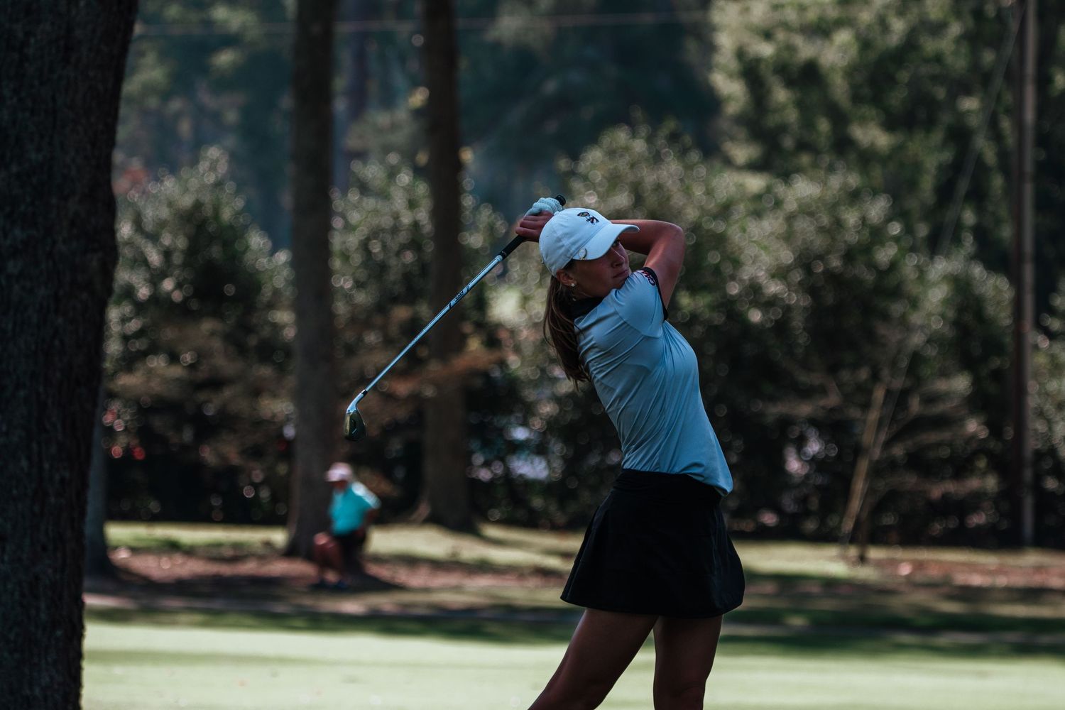 Golfer in light blue attire swings a club on a tree-lined course.