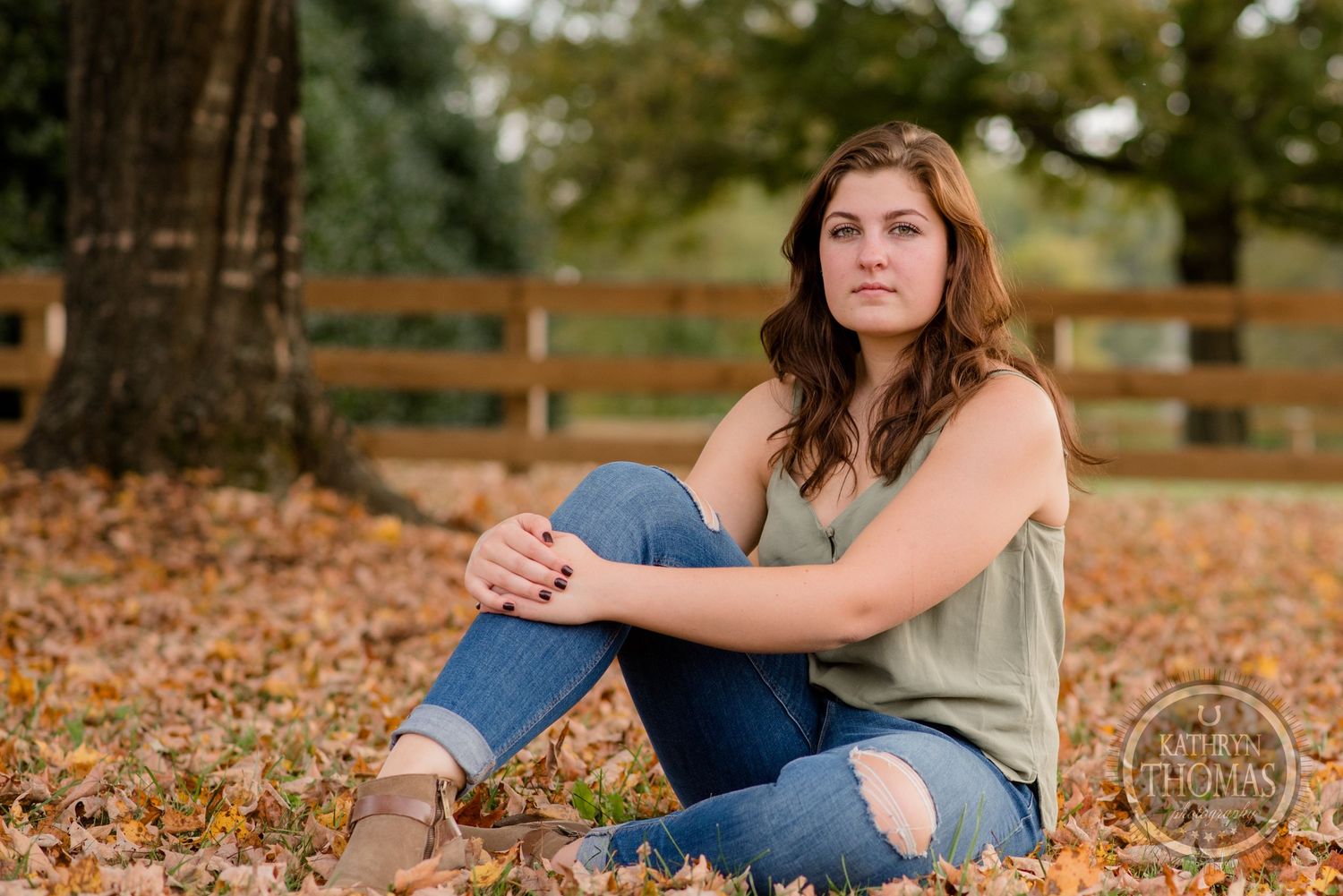 Casual portrait sitting among fall leaves with wooden fence and autumn trees in background.