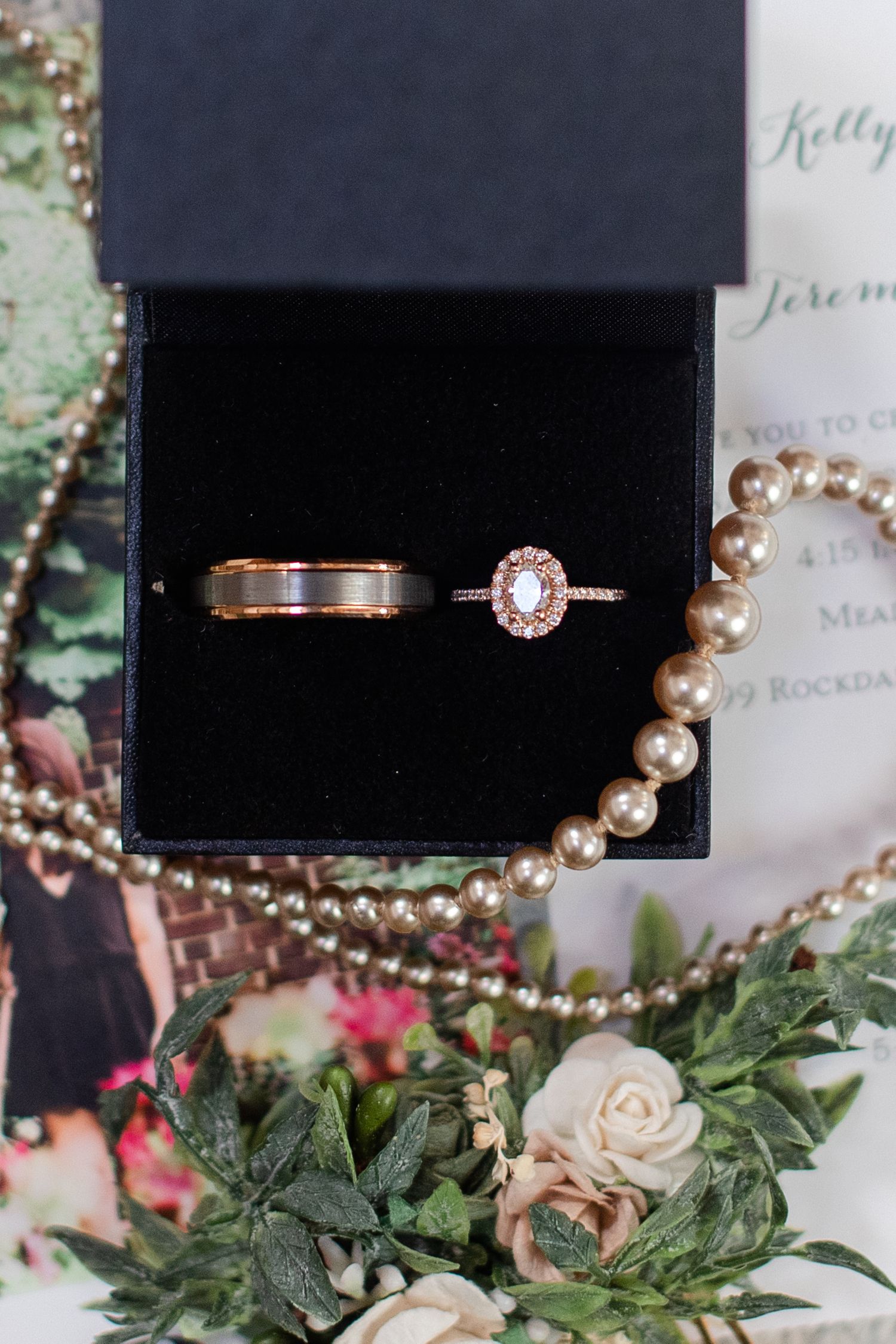 Wedding rings and jewelry displayed with blush roses and greenery on invitation paper.