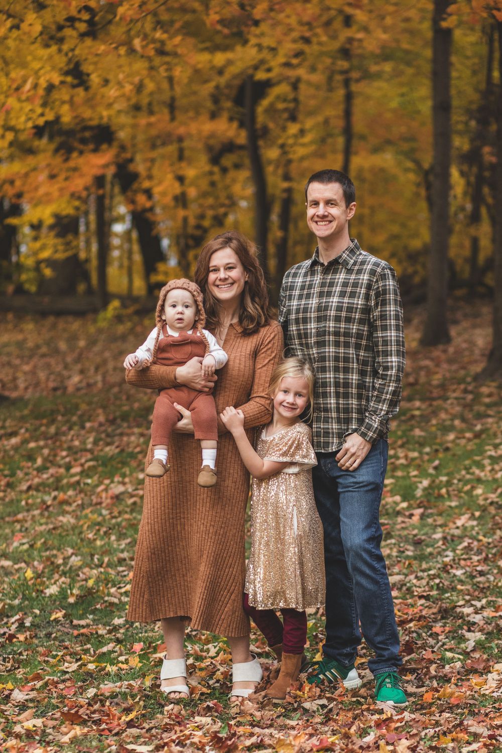 A family of four poses together among fallen autumn leaves in a park.