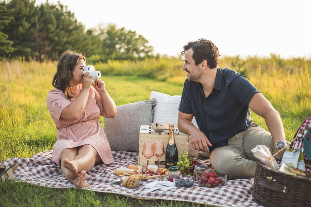 A couple enjoys a romantic picnic with wine and snacks on a blanket in a sunny field.