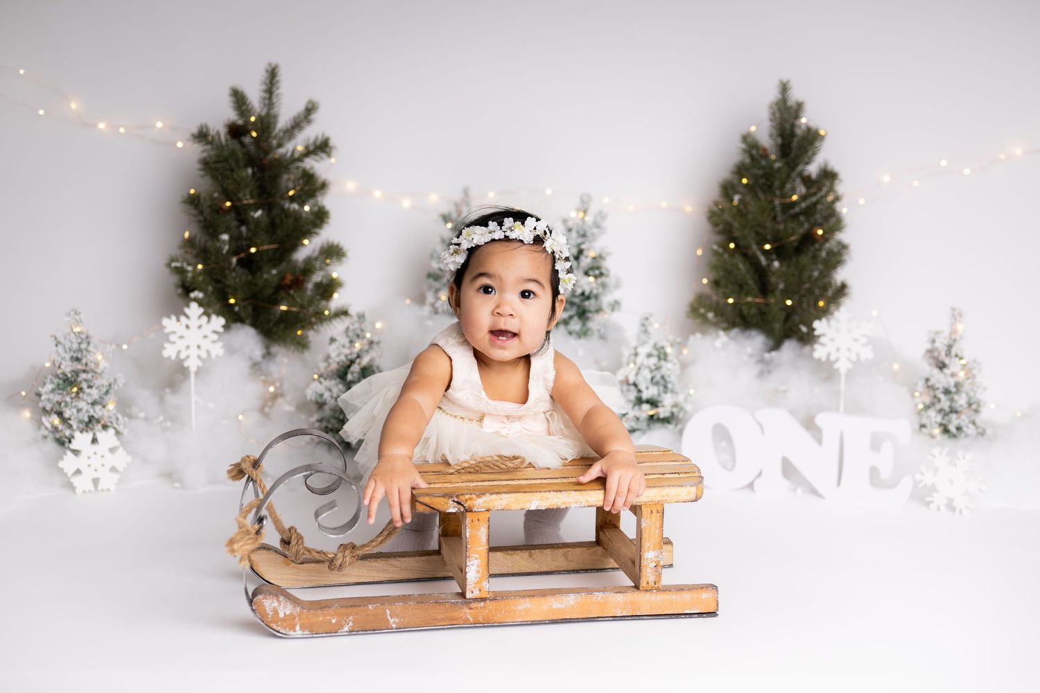 A vintage wooden sled sits in a winter wonderland scene with decorated evergreen trees and string lights.