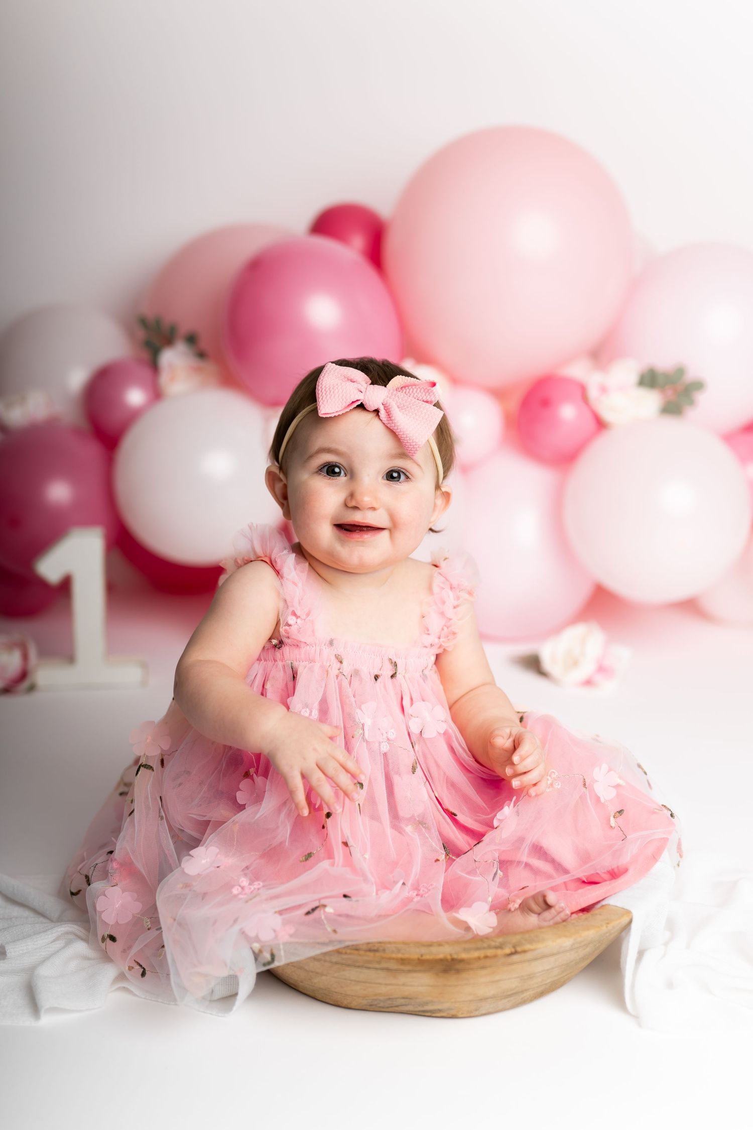 Baby in pink tulle dress sitting on wooden platform with pink balloon arch backdrop.
