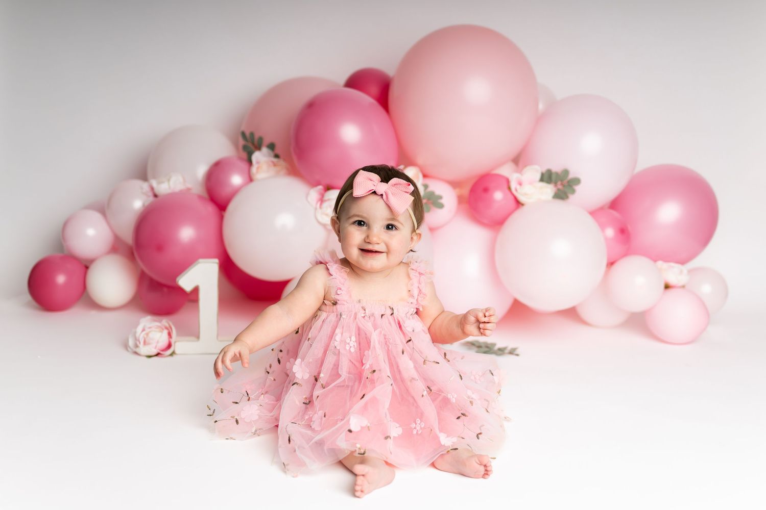 Baby standing in pink tulle dress against pink balloon backdrop for first birthday.