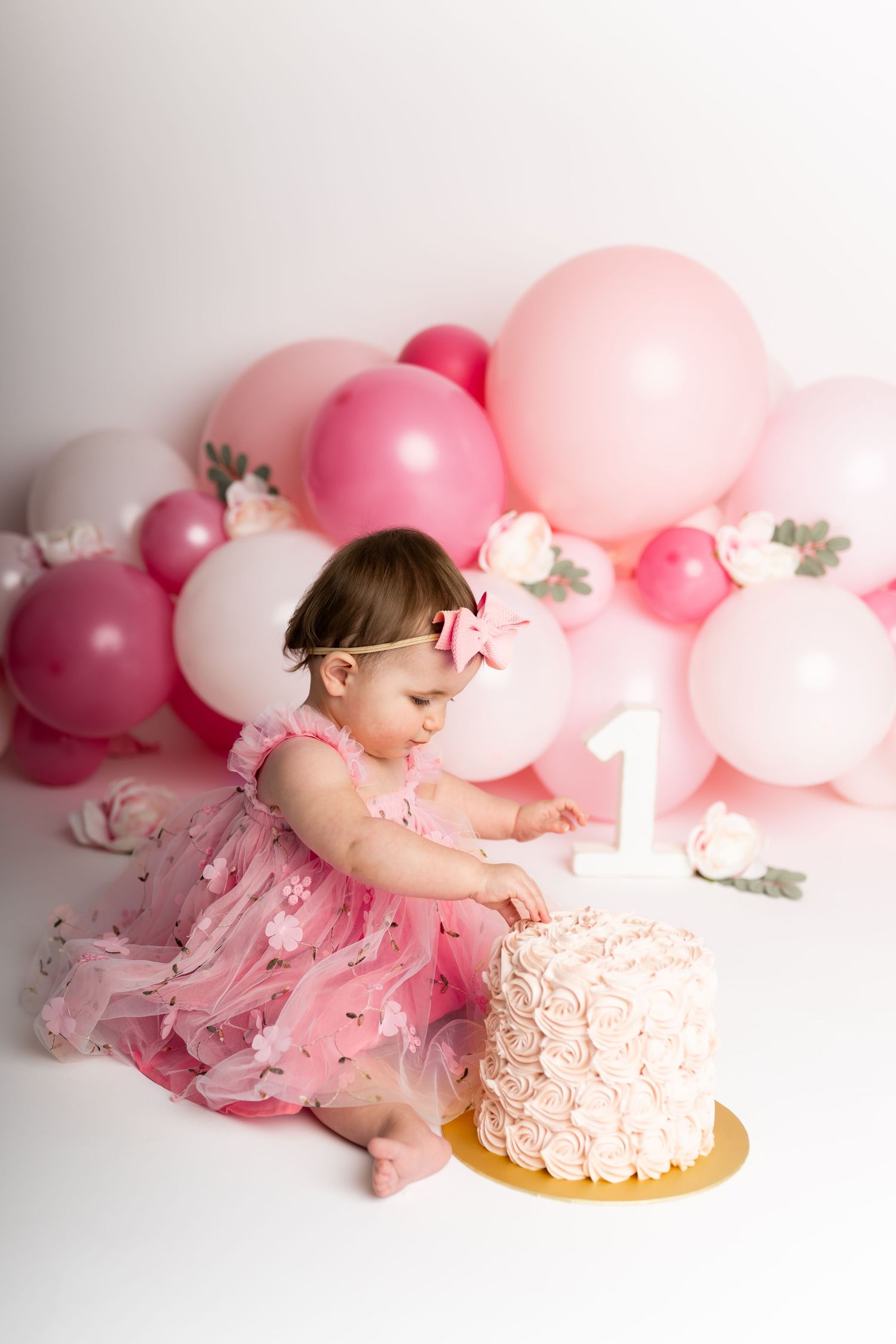 Baby in pink dress reaching for first birthday cake with balloon arch decoration.