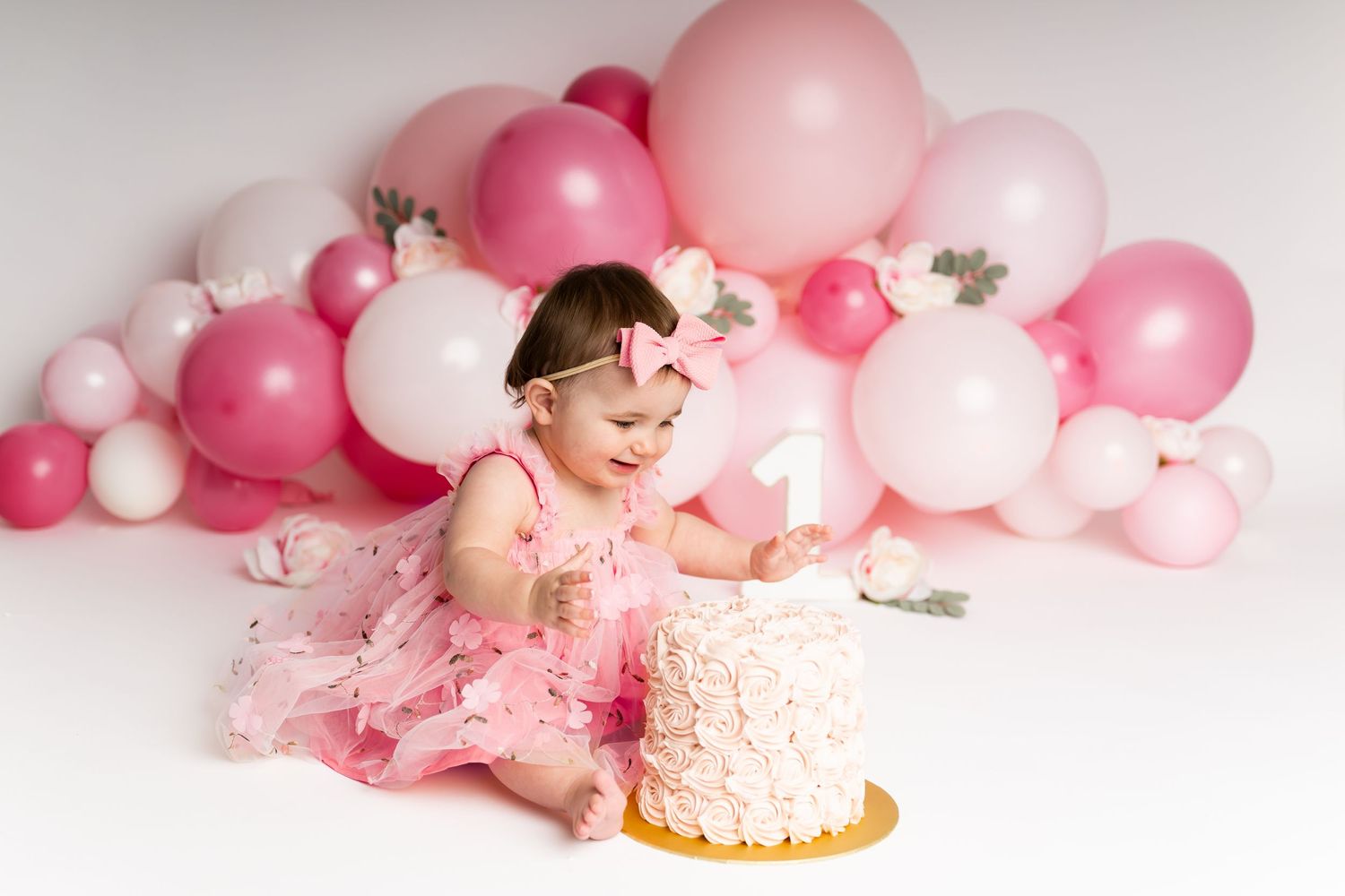 Baby enjoying first birthday cake smash with pink balloon backdrop.