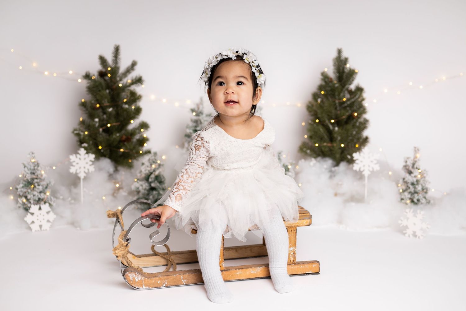 A winter wonderland photo setup with small evergreen trees, string lights and a wooden sled against a white backdrop.