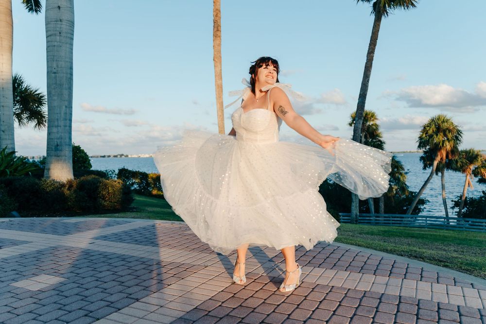 Bride in flowing white dress twirls playfully on a waterfront walkway at sunset.