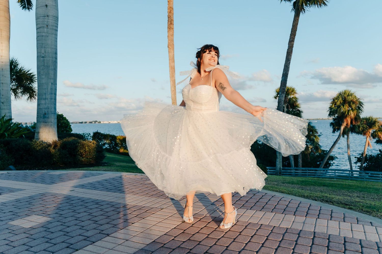 Bride in flowing white dress twirls playfully on a waterfront walkway at sunset.