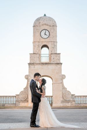 A couple in wedding attire embraces in front of historic clock tower at sunset.