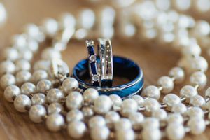 Wedding rings and pearl necklace arranged on blue surface for detail photo.