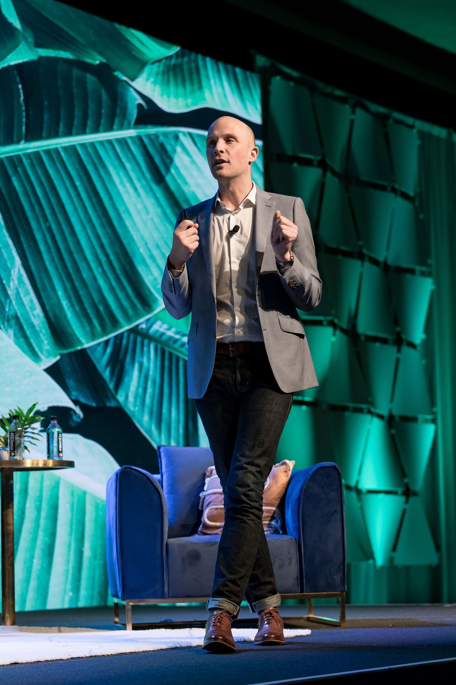 A speaker presents on a stage with dramatic turquoise lighting and curtains in the background.