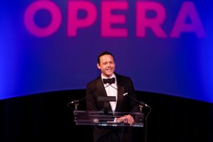 Speaker at podium with Opera sign illuminated in bright pink against dark blue background.