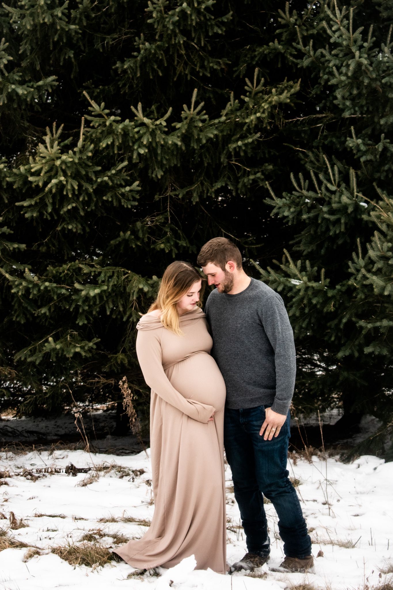 A winter maternity photoshoot with a couple standing together against evergreen trees in snow.