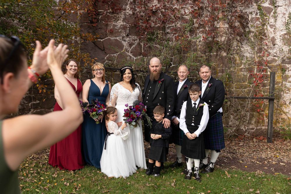 Wedding party photo being taken in front of rustic brick wall with traditional Scottish attire.