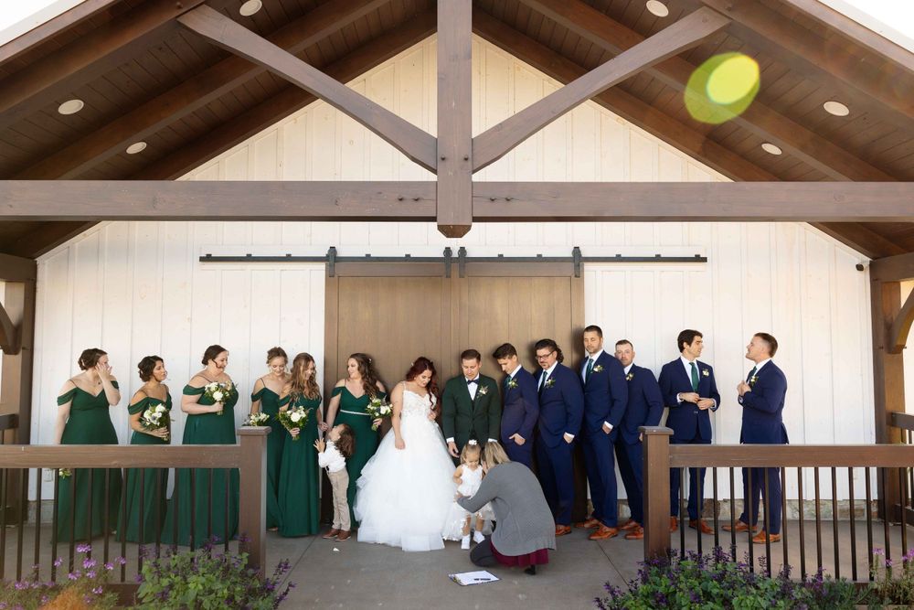 Wedding party in emerald green and navy attire posed under wooden beamed pavilion.
