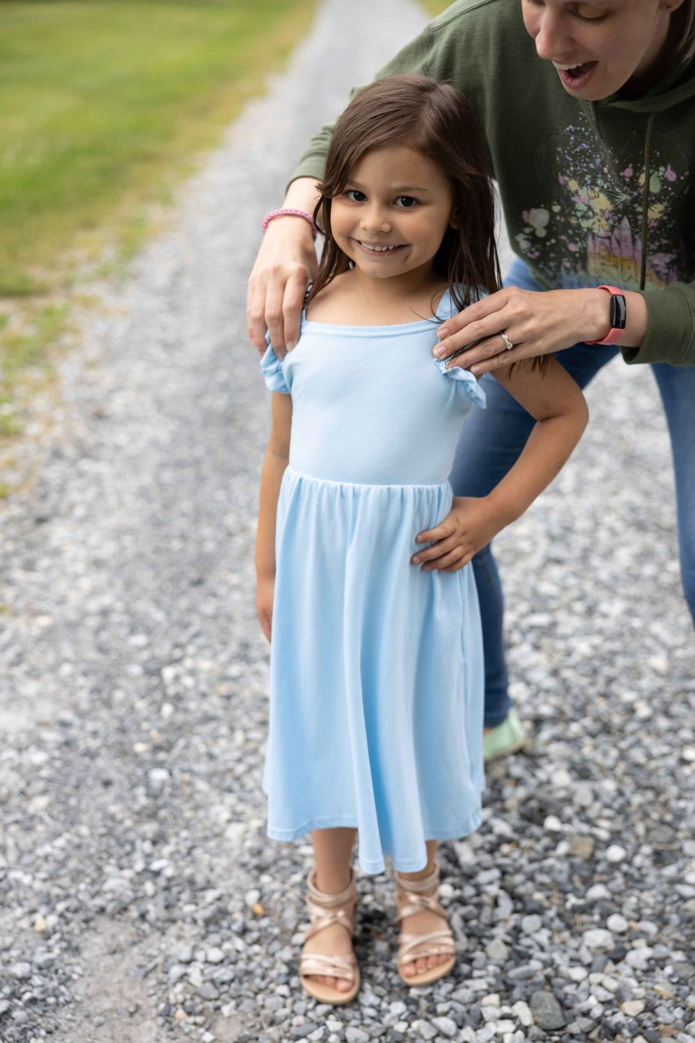 A child in a light blue dress stands on a gravel path with hands on hips.