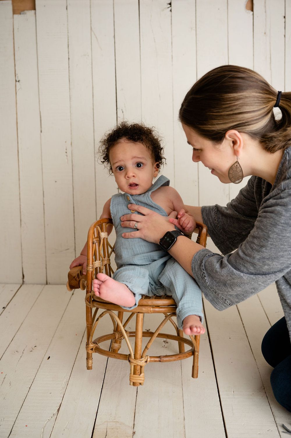 A sweet moment captured with a small child seated in a rattan chair against a white wooden backdrop.