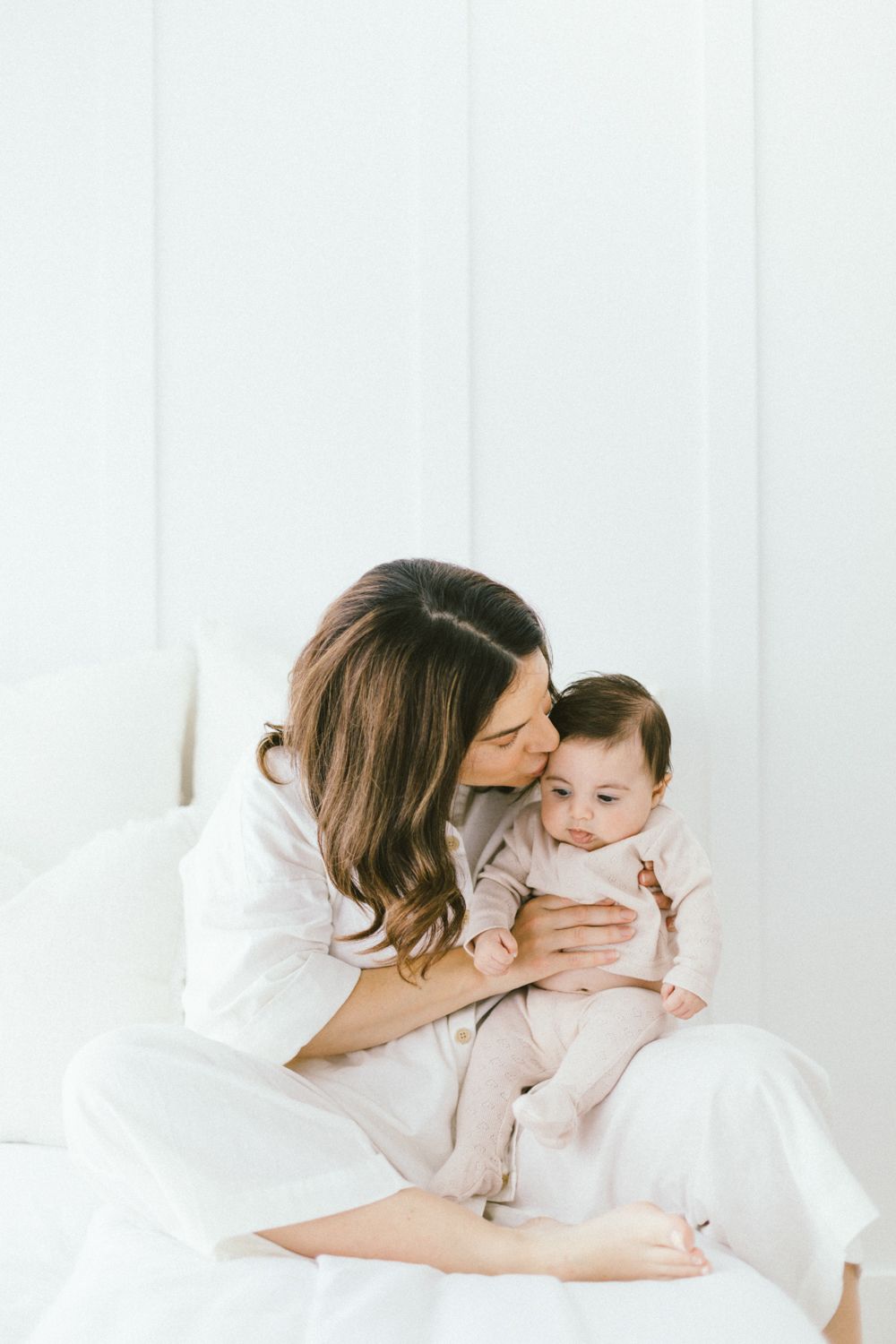 Tender moment between caregiver and infant wearing light colored clothing against white background.