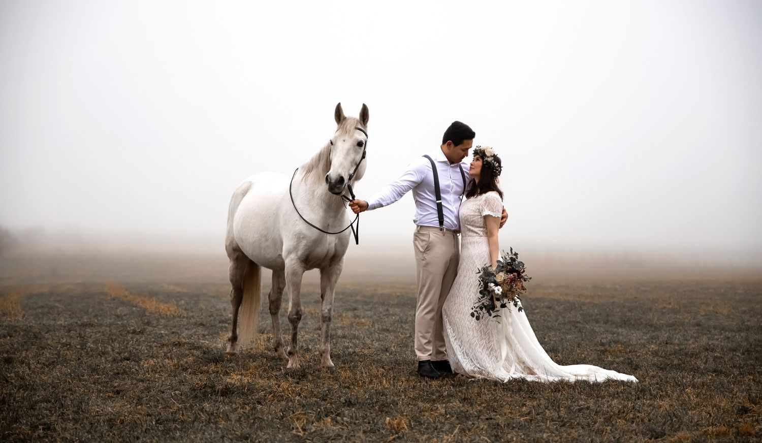 Misty wedding photo with couple and white horse in rustic outdoor setting.