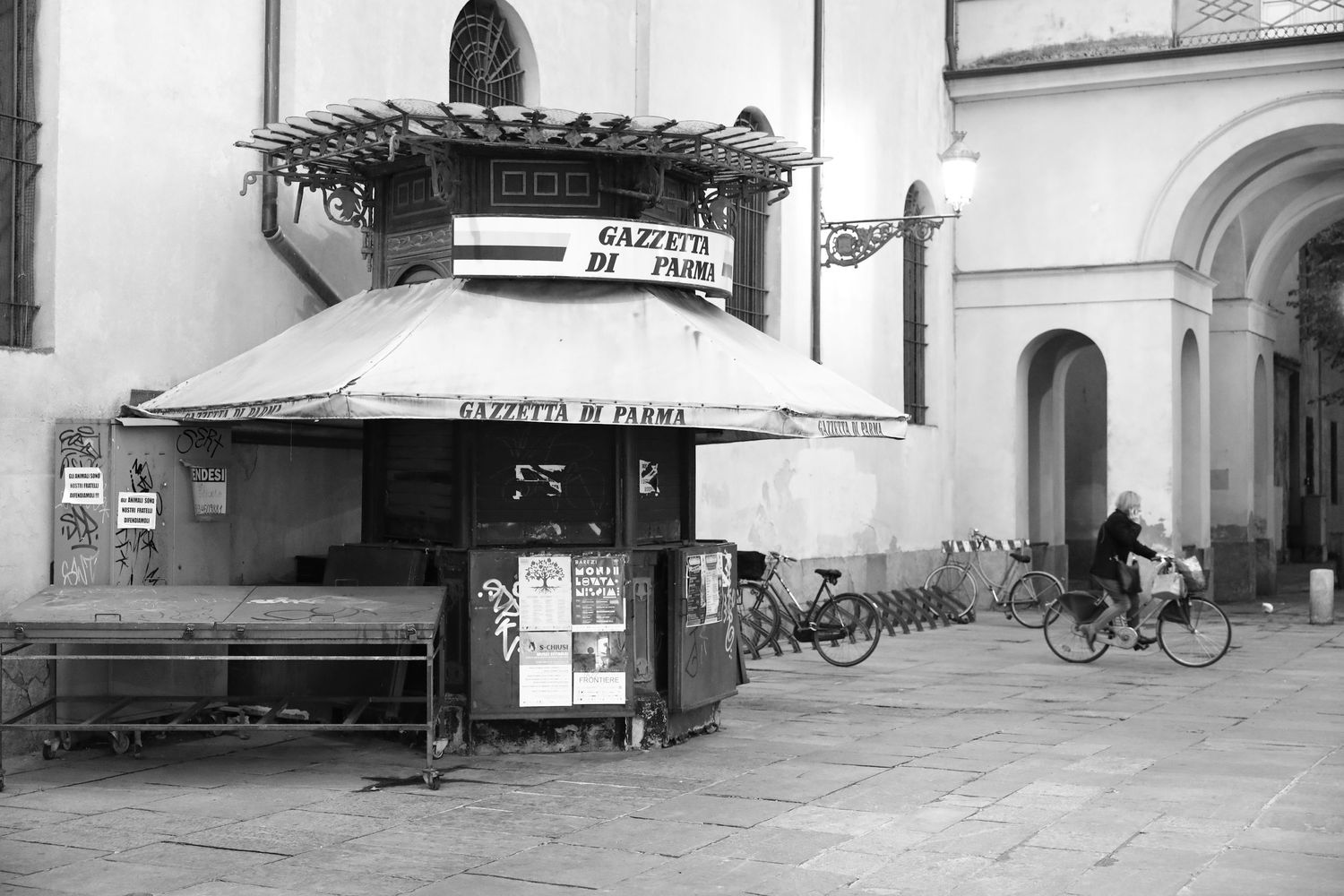 Black and white photo of a vintage newsstand with bicycles parked nearby in an Italian city.