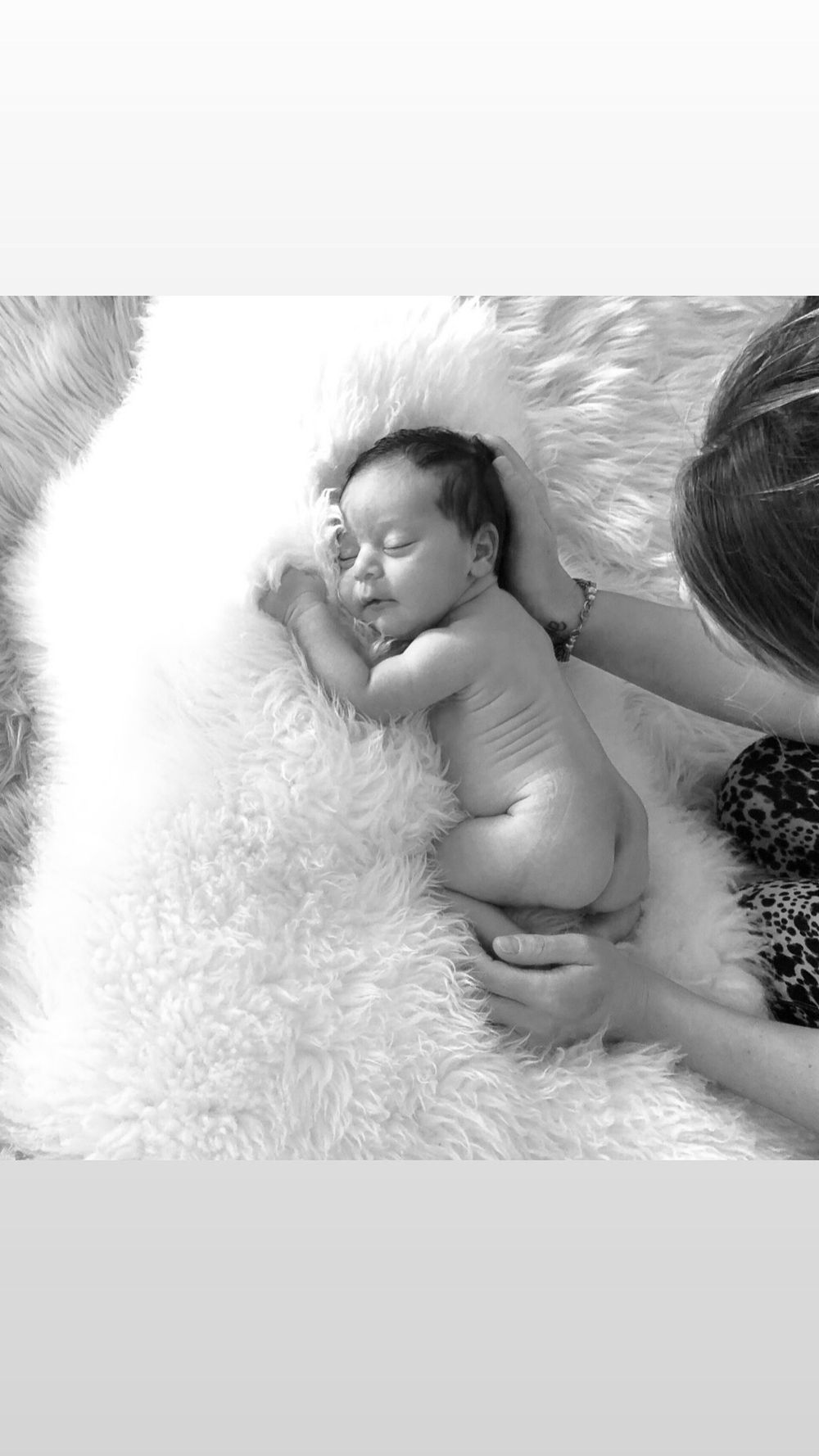 Small baby lying peacefully on soft white fur blanket in black and white photograph.